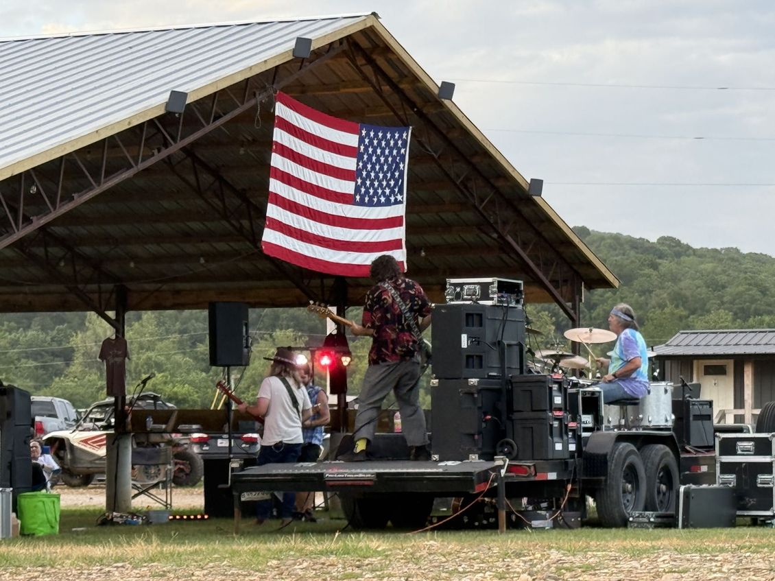 A band performs on a trailer stage under a pavilion featuring an American flag, with trees and buildings in the background.