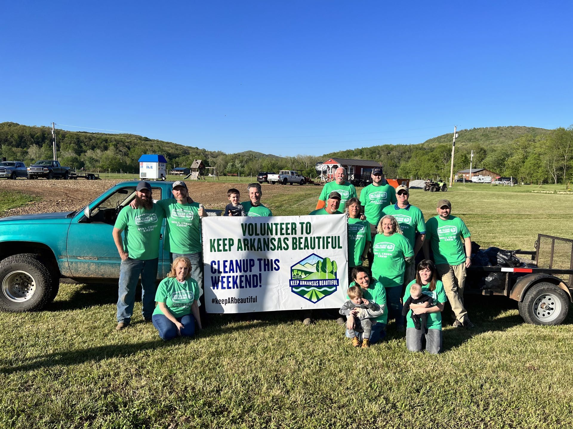 A diverse group of people stands in a grassy field next to a truck, holding a banner promoting a local cleanup event.