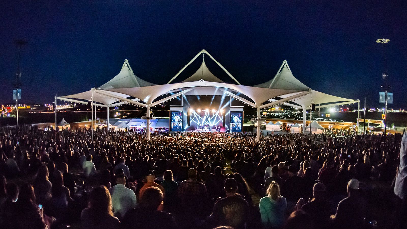 Large outdoor concert at night with a brightly lit stage and a dense crowd under white canopy tents