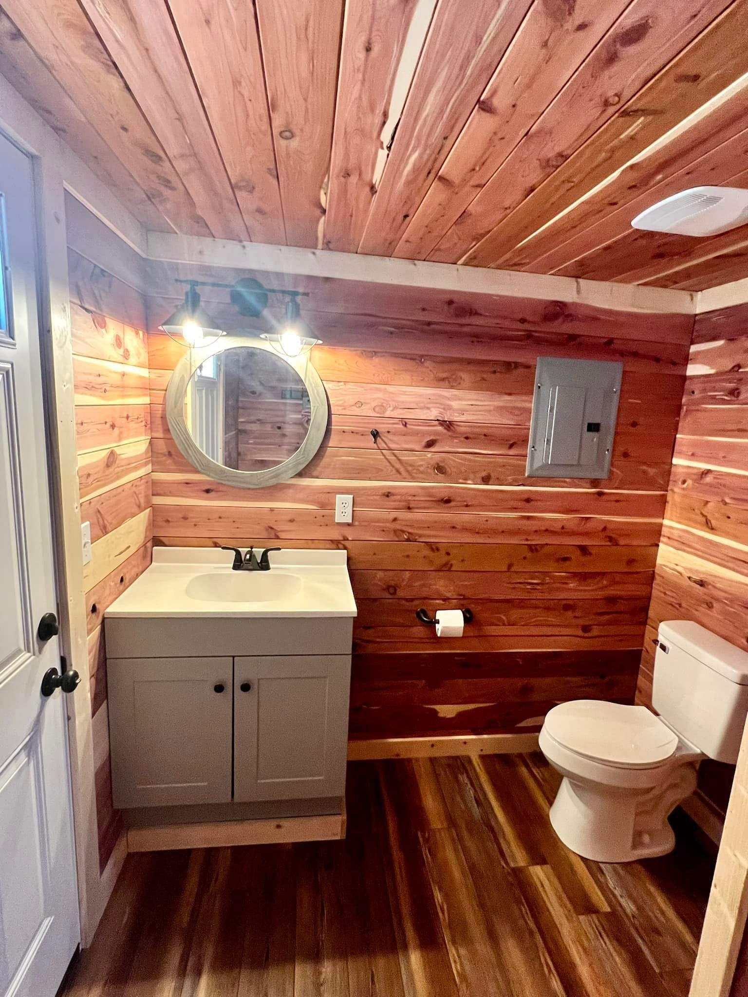 Rustic bathroom with wood walls and ceiling, vanity, round mirror, toilet, and shower door.