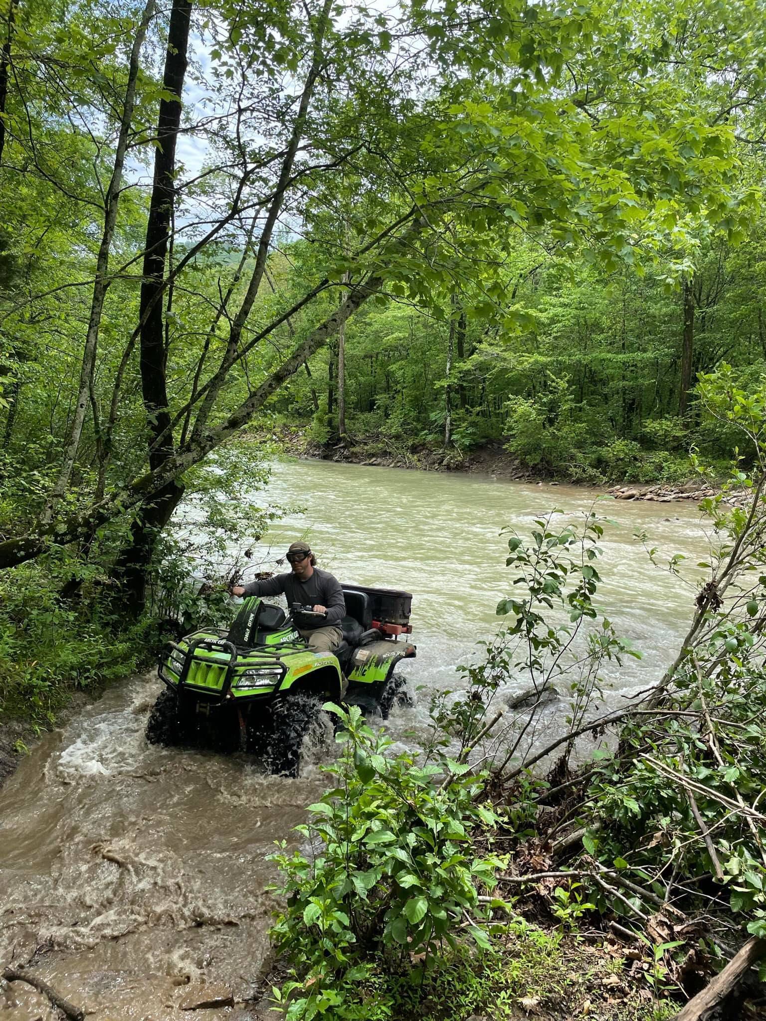 A person rides a bright green ATV through a shallow, muddy river surrounded by lush green forest.