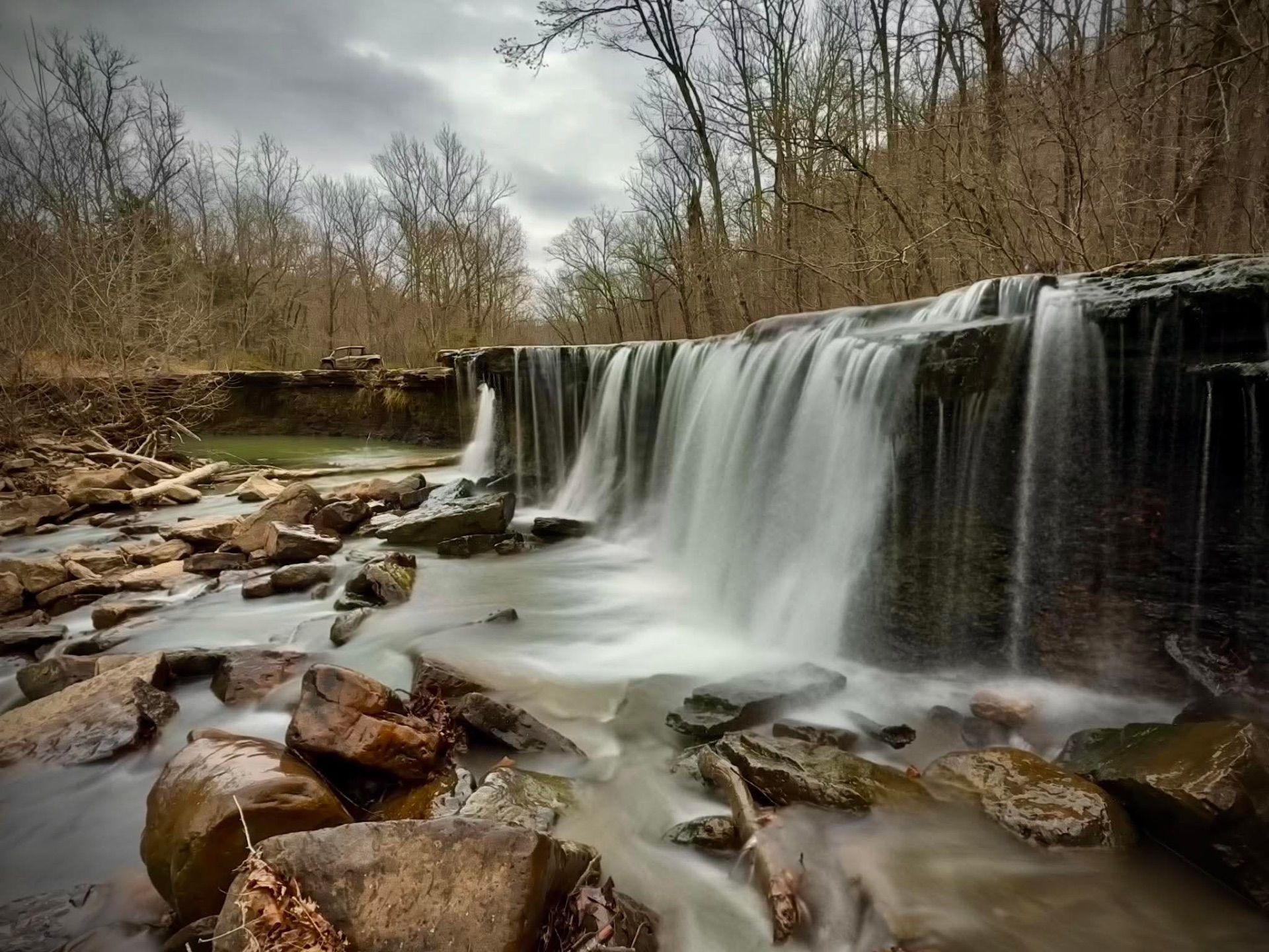 A wide, tiered waterfall flows over a mossy rock ledge into a calm pool surrounded by bare trees under a cloudy sky.