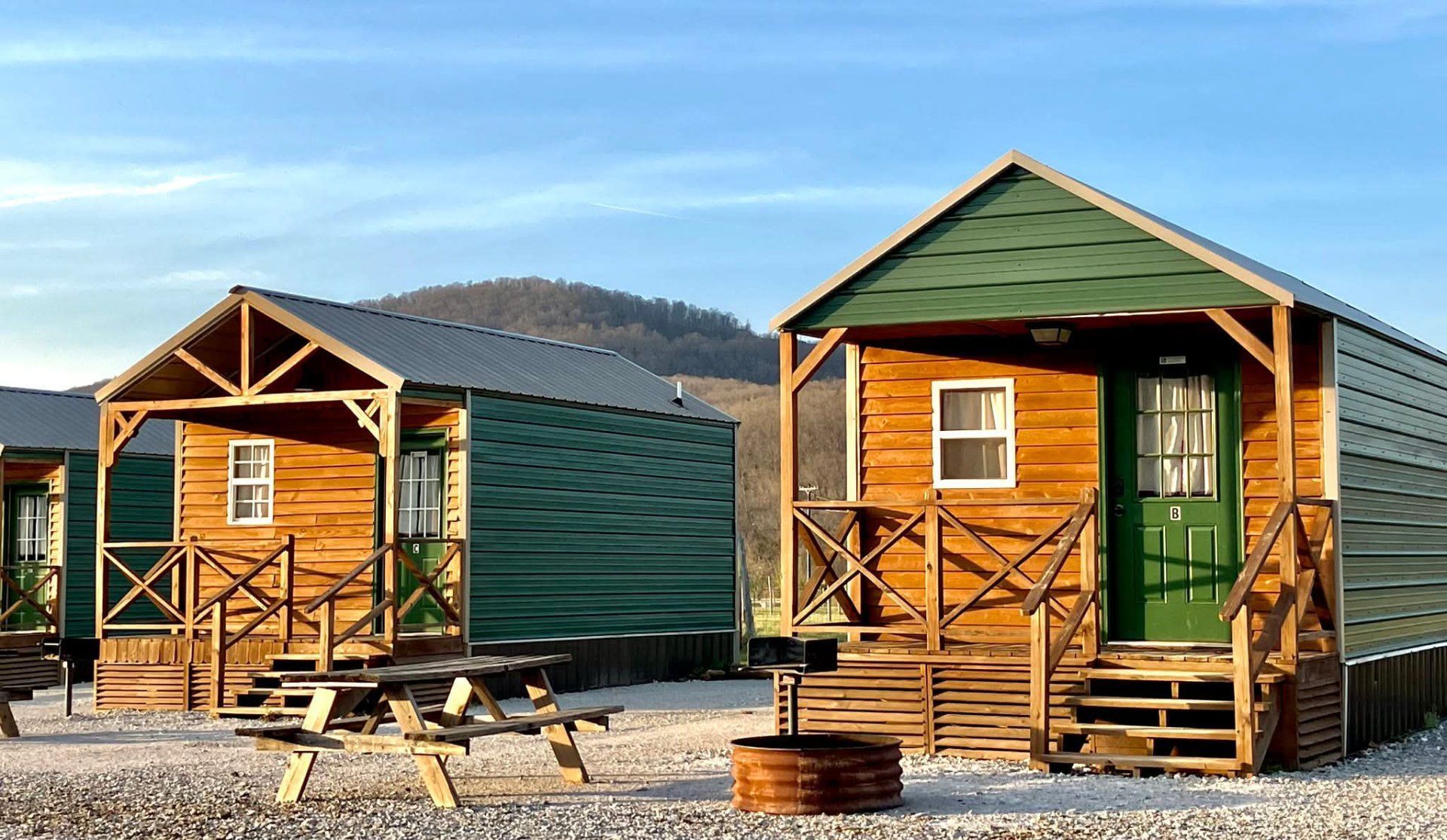 Two small wooden cabins with green trim and porches stand on a gravel campsite against a mountain backdrop.