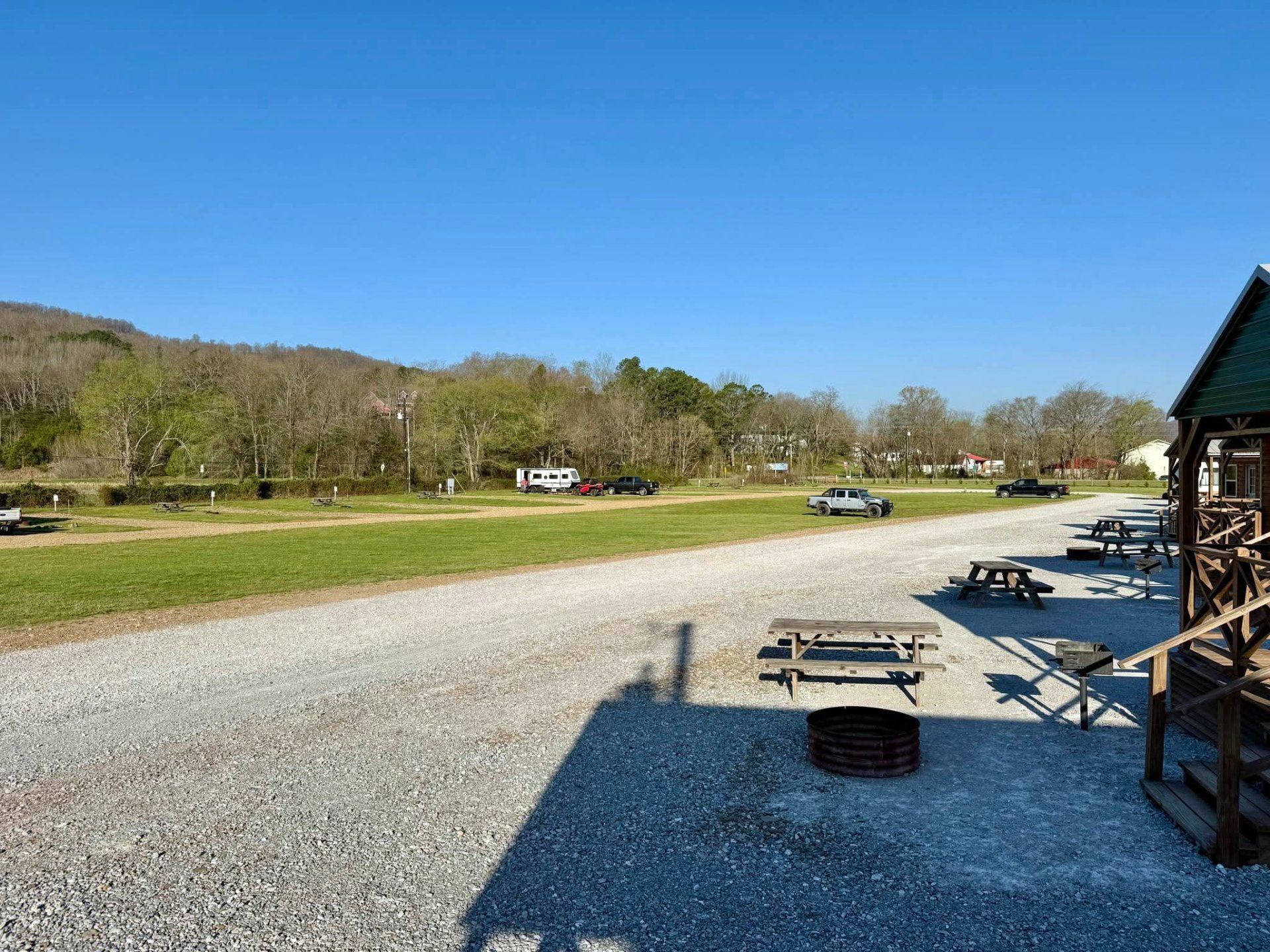 An outdoor campground area with picnic tables and a fire pit on a gravel lot under a clear blue sky.