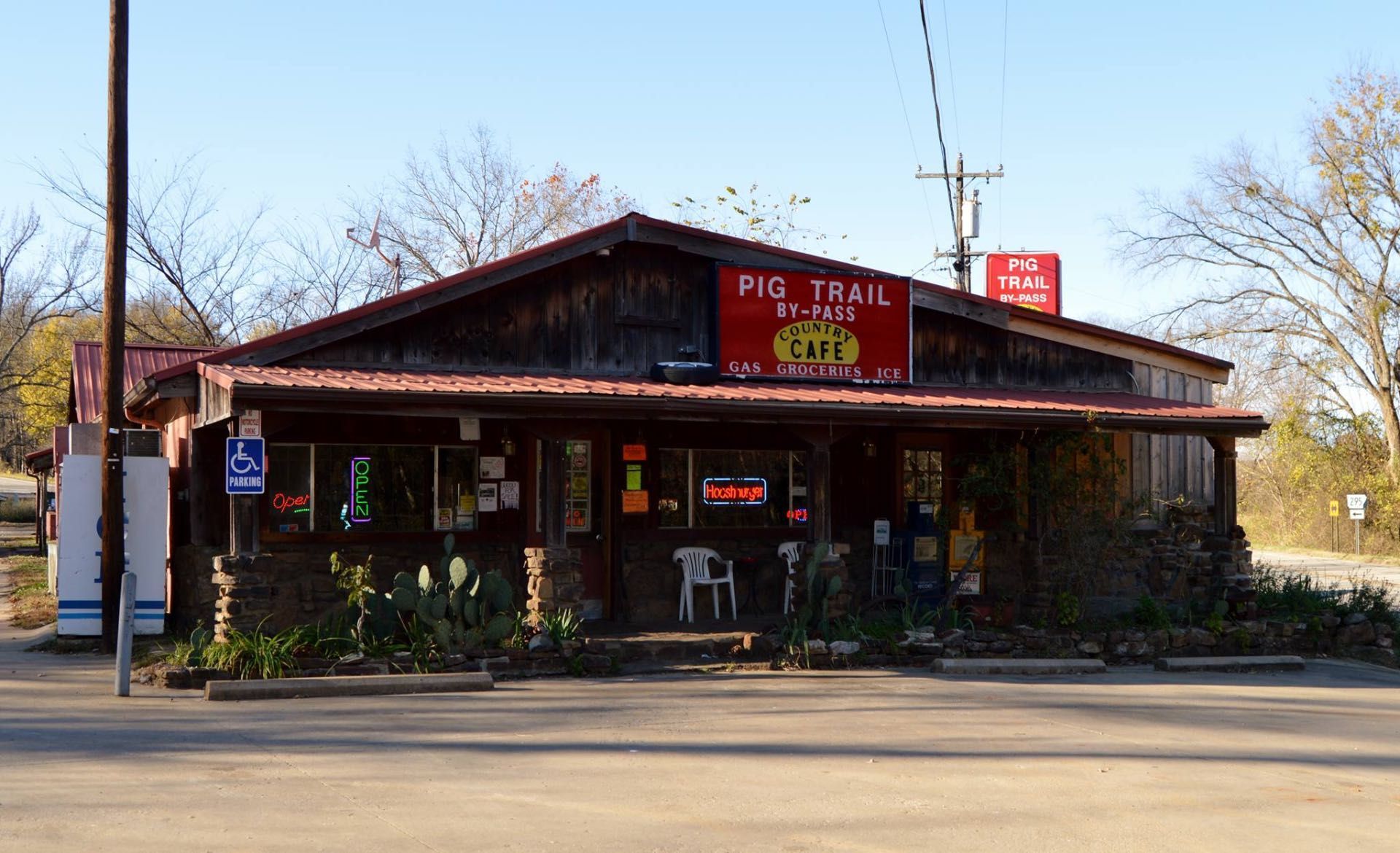 Pig Trail By-Pass Cafe, a rustic wooden building with a red sign, stone base, and outdoor seating on a sunny day.