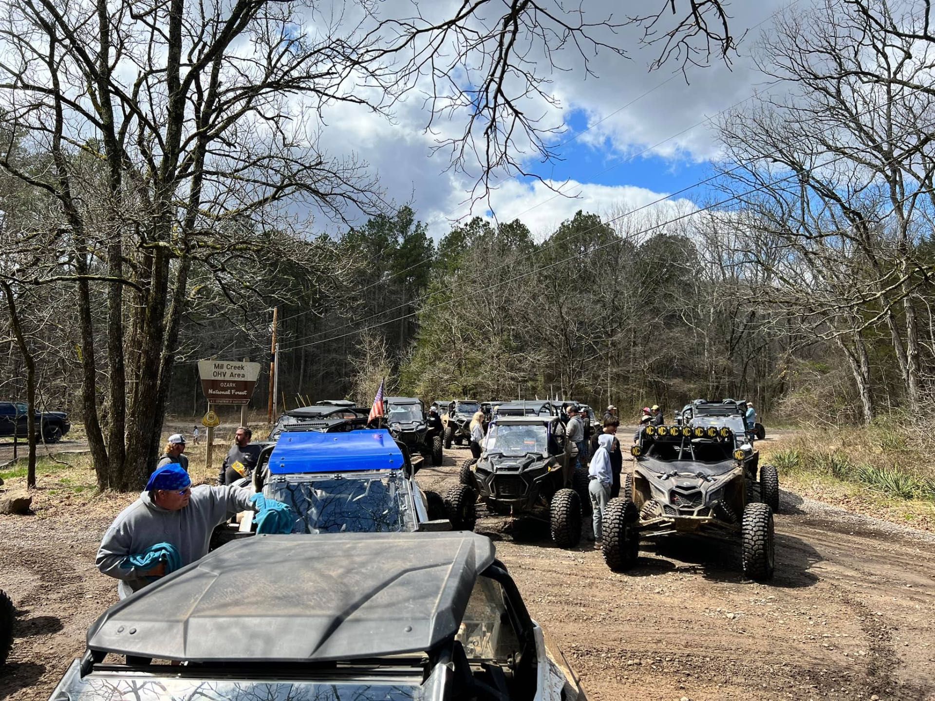 A group of off-road vehicles parked on a dirt trail in a forest on a sunny day, with a person standing by a blue UTV.