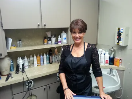 Woman in salon apron smiles next to a sink, surrounded by hair products.