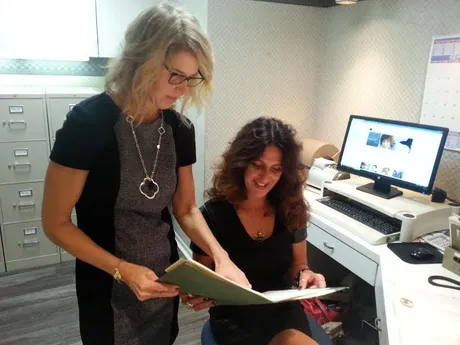 Two women looking at a file in an office setting, one standing, the other seated, near a computer.