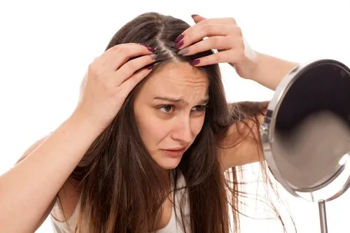 Woman examining her scalp for hair loss, looking concerned in front of a mirror.