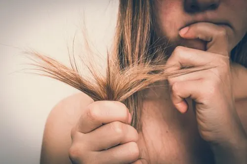 Woman holding split ends of hair, biting nails, close-up, neutral background.