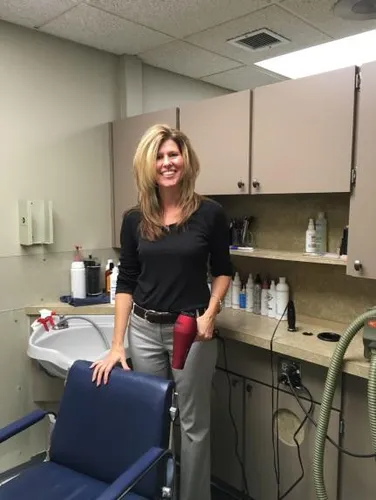 Woman in a salon, holding a hair dryer. She smiles and stands near a styling chair and sink.