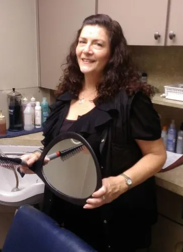 Woman holding a mirror in a salon. She smiles, surrounded by beauty products and a sink.