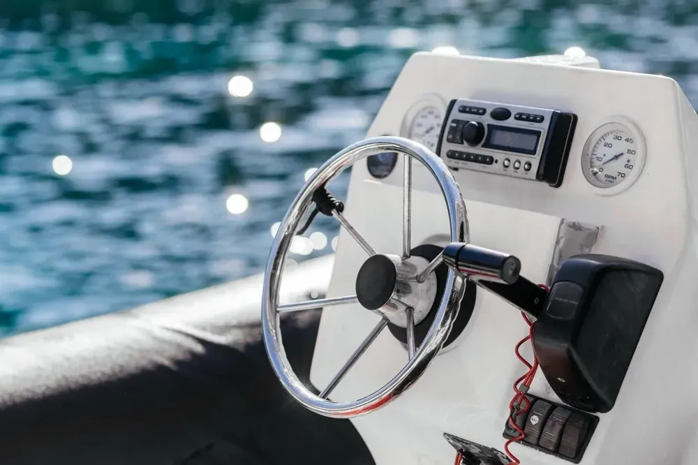 The control console of a boat with a chrome steering wheel, a radio, and gauges, positioned against blurry open water. — Ocean Mate Marine Services in Coomera, QLD