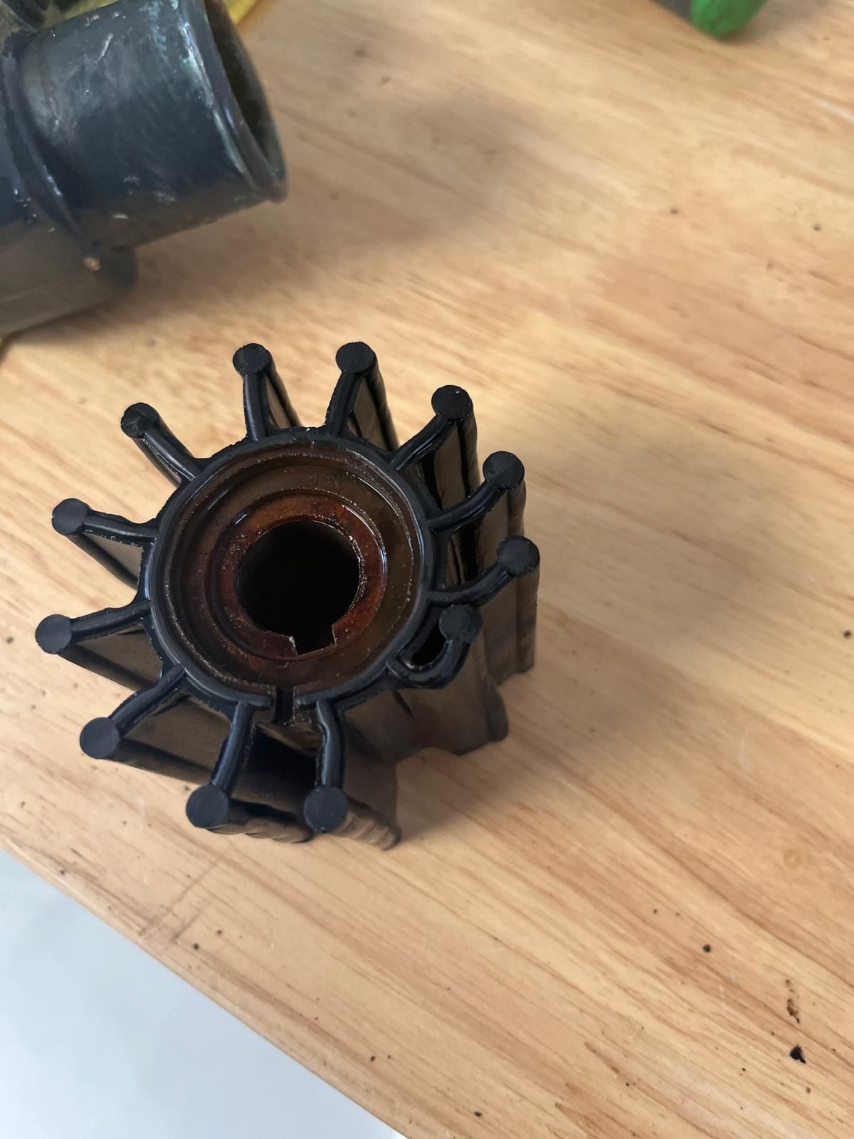 A black rubber water pump impeller with twelve vanes sitting on a wooden surface next to a metal housing part. — Ocean Mate Marine Services in Coomera, QLD 