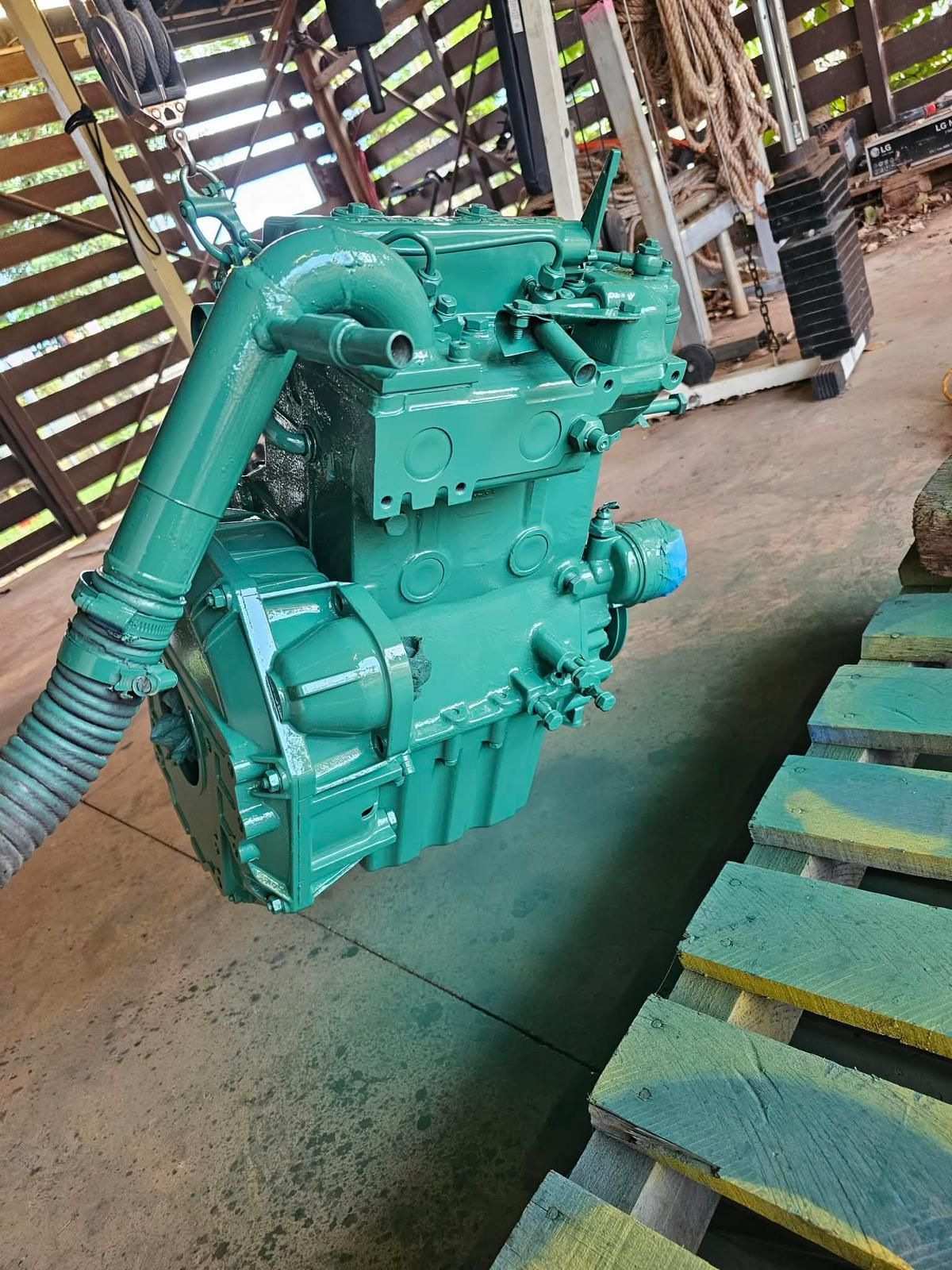 A bright green industrial engine hangs suspended by a chain hoist in a rustic wooden building near a wooden pallet. — Ocean Mate Marine Services in Brisbane, QLD