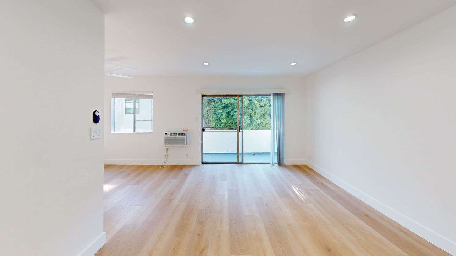 Empty room with hardwood floors, white walls, and a sliding glass door to a balcony with trees.