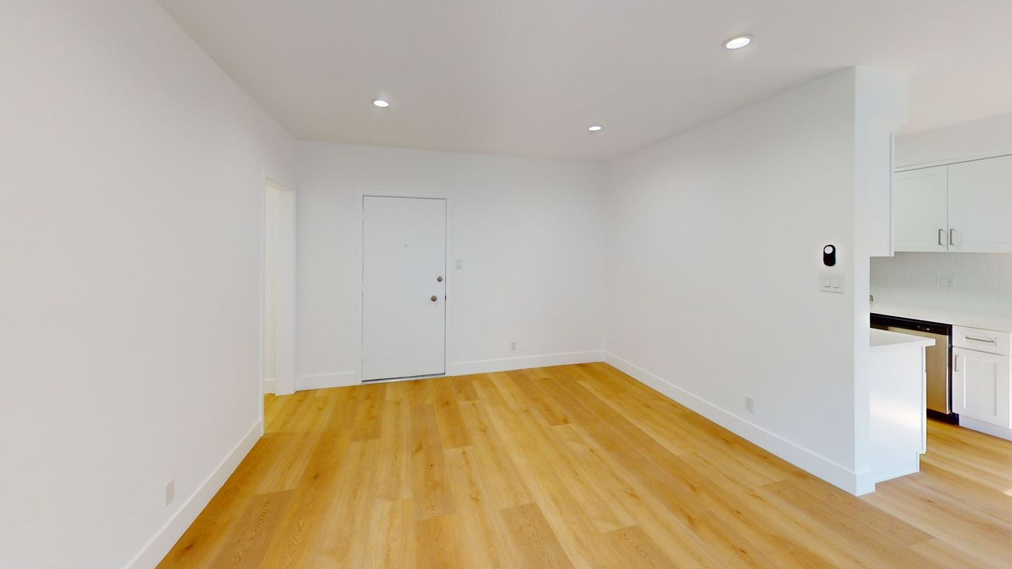 Empty room with light wood floors, white walls, and recessed lighting. A door and cabinets are visible.