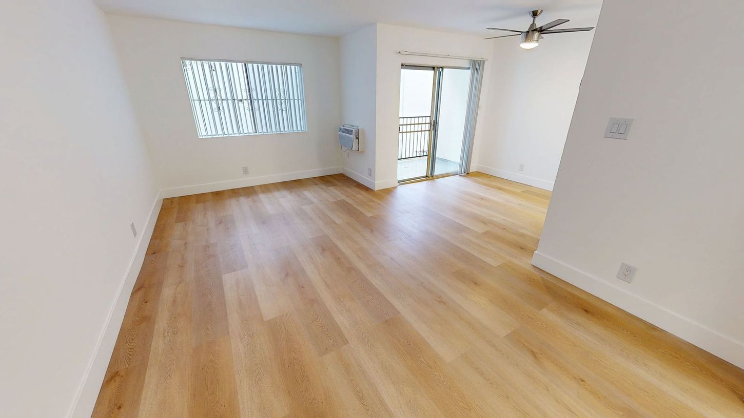 Empty room with light wood floors, a window, and a sliding glass door to a balcony. White walls.