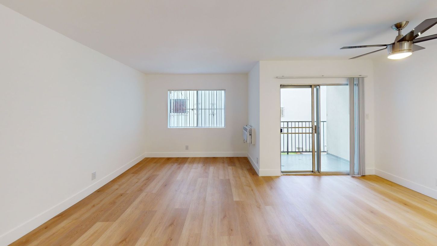Empty apartment interior with wooden floors, white walls, sliding door to balcony, and a ceiling fan.