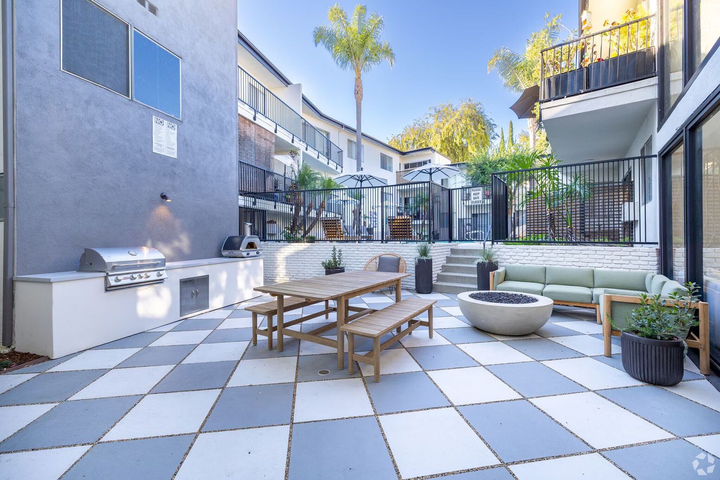 Patio with grill, seating, dining table, and checkered floor. Apartment building in background. Sunny day.