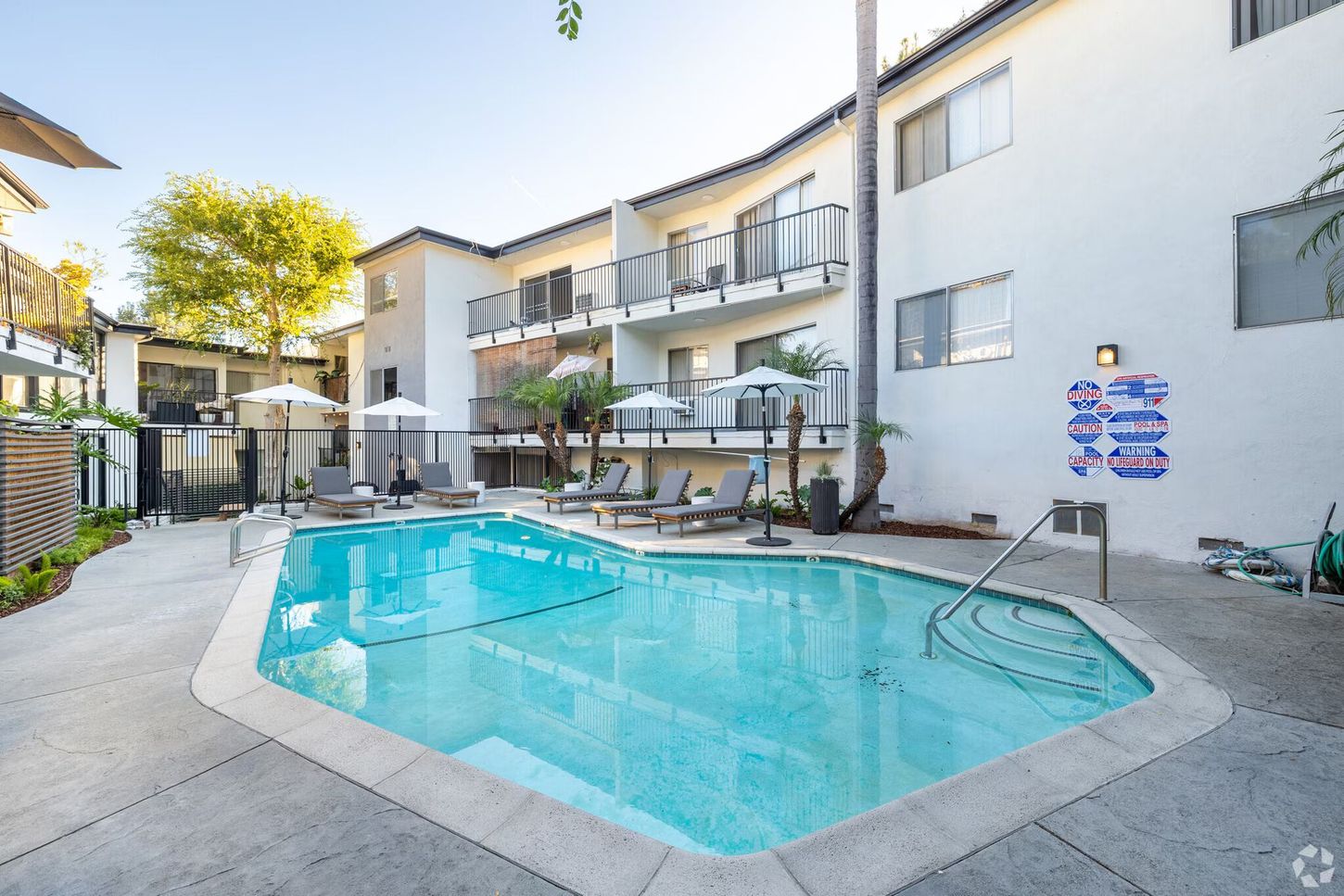 Pool and apartment buildings with lounge chairs, umbrellas, and palm trees.