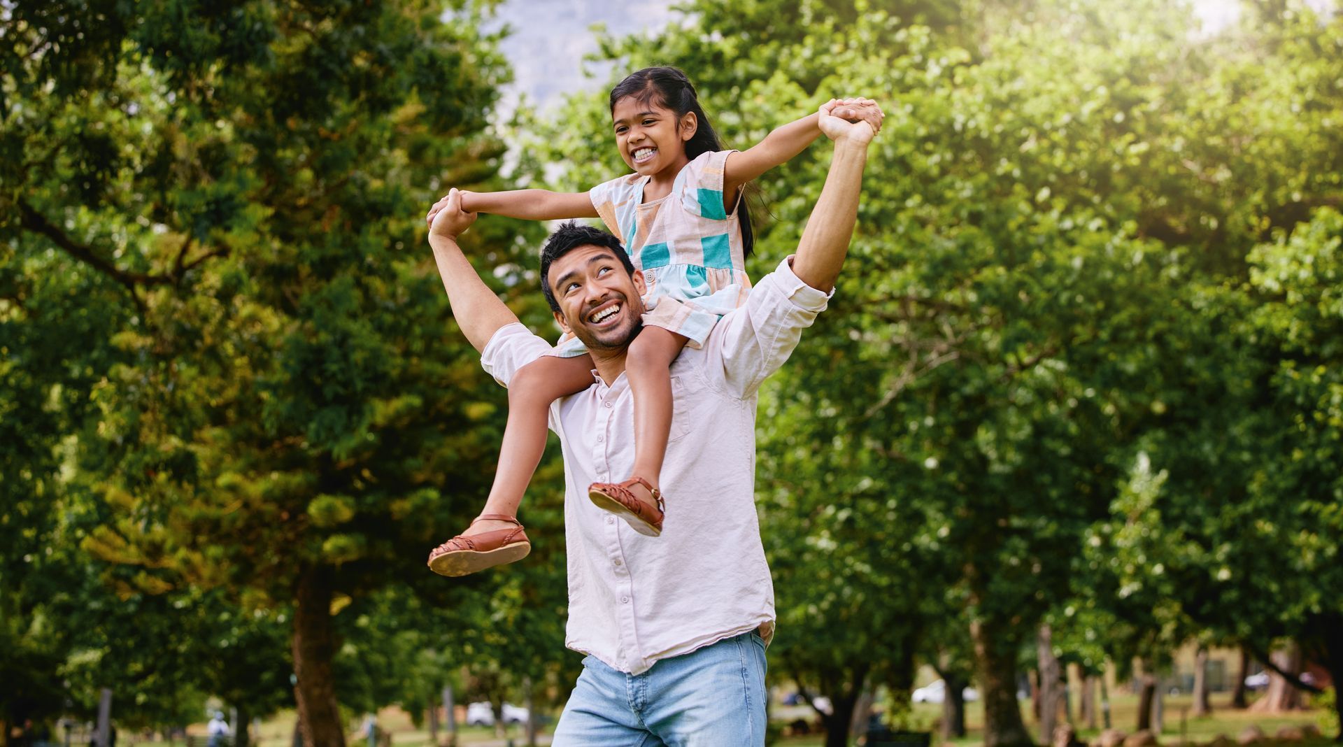 A smiling father giving his young daughter a shoulder ride in a sunny park. The little girl laughs while holding her arms up in the air, surrounded by lush green trees under bright daylight.