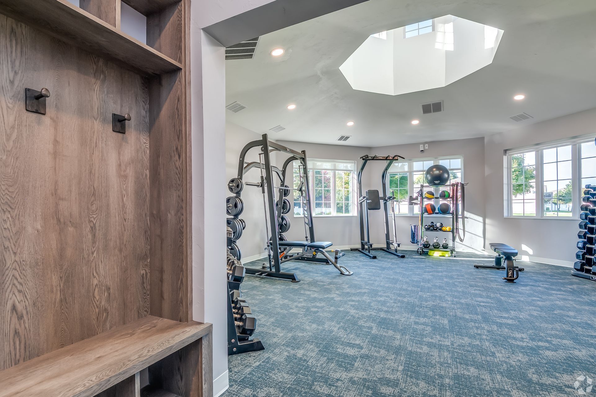 Bright gym room with workout equipment, blue carpet, and large windows.