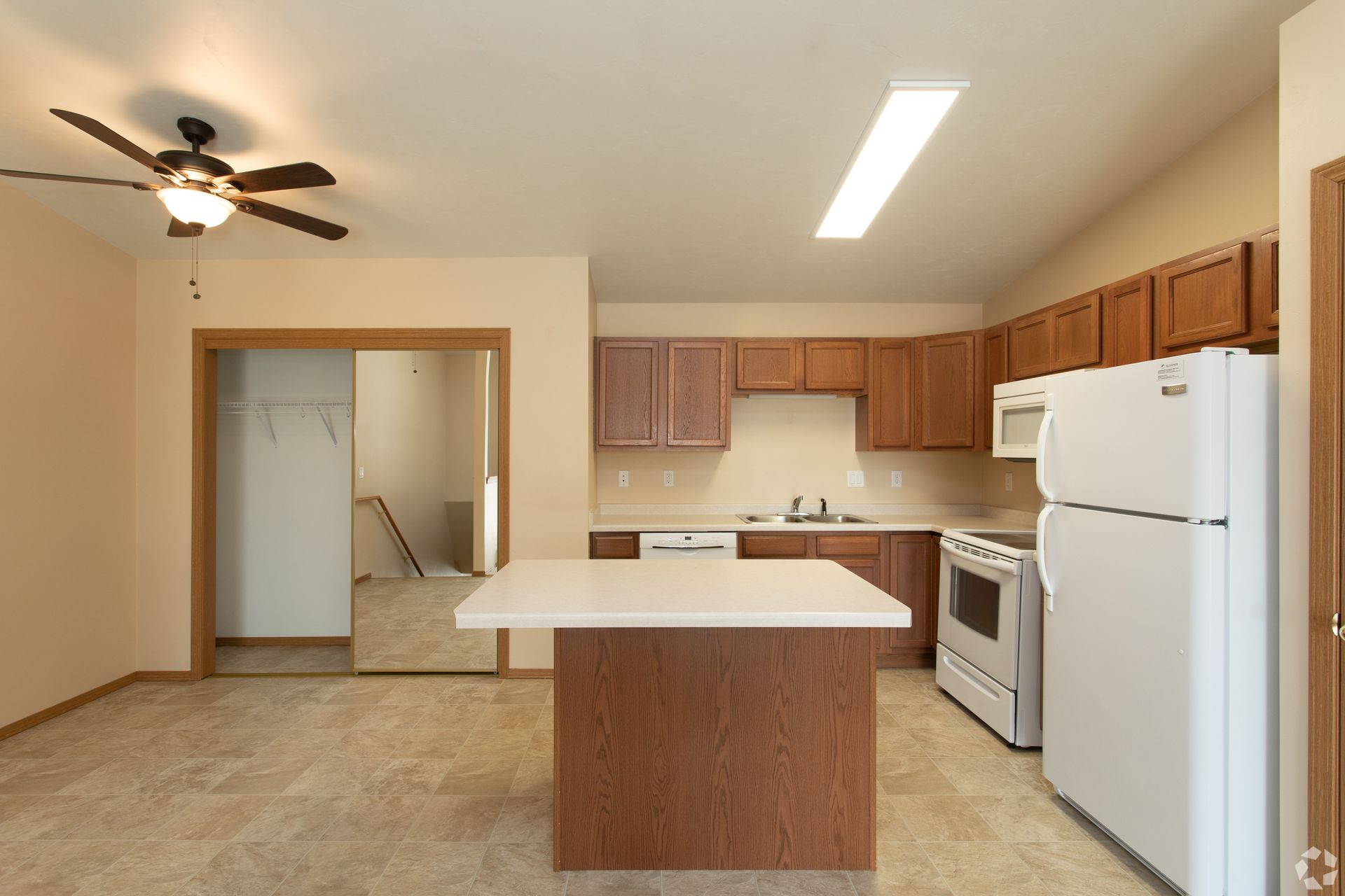 Empty kitchen with wood cabinets, white appliances, and a center island in beige tile room