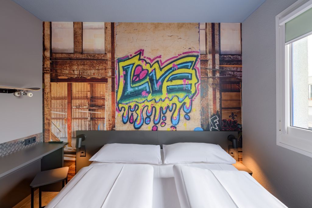 A hotel room with a white bed in front of a wall mural featuring graffiti that says 