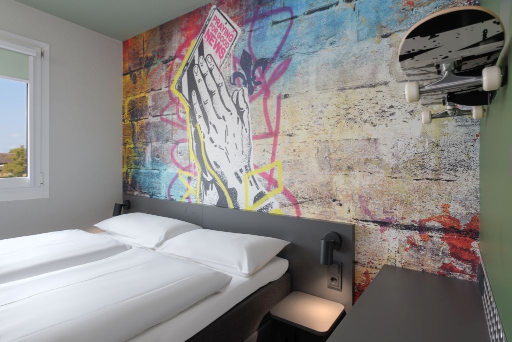 A modern bedroom featuring a street-art mural of praying hands behind a bed with white bedding and a wall-mounted skateboard.