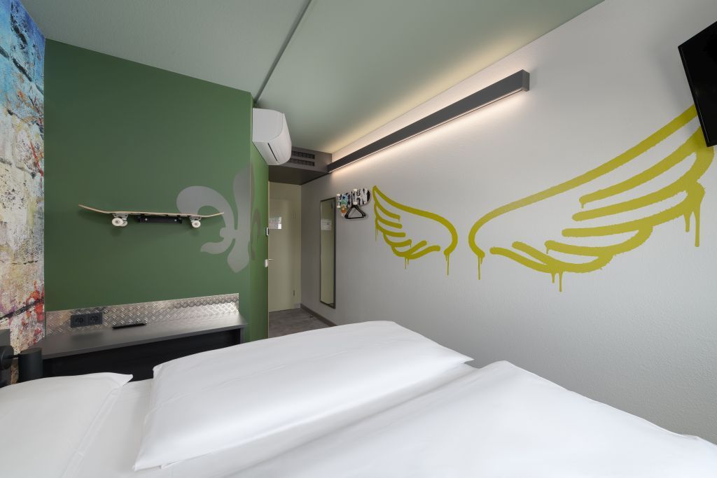 A modern hotel room with a green wall, a white bed, and yellow graffiti-style wings painted on the white wall.