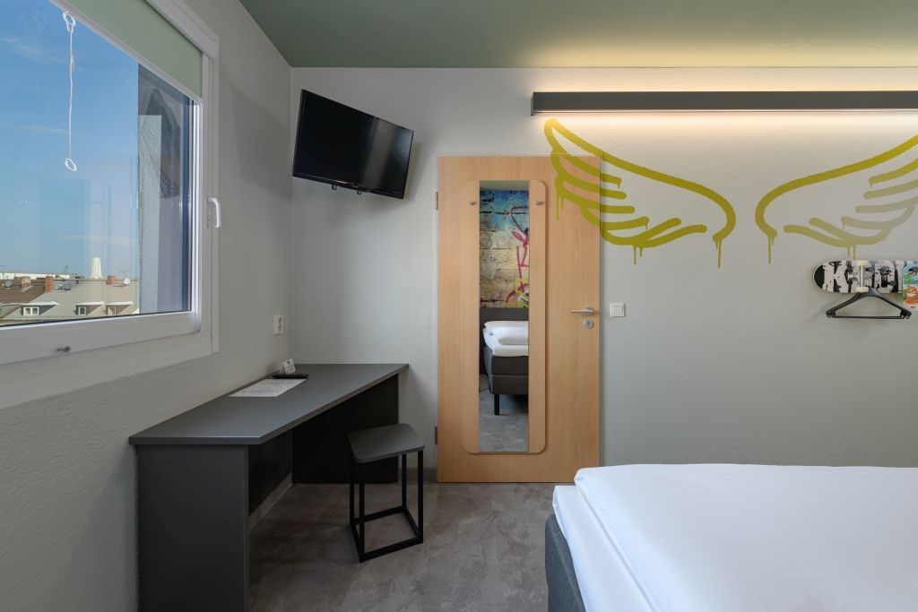 A hotel room with a white bed, a desk, a TV, a mirror on a wooden door, and gold angel wings mural on a light gray wall.