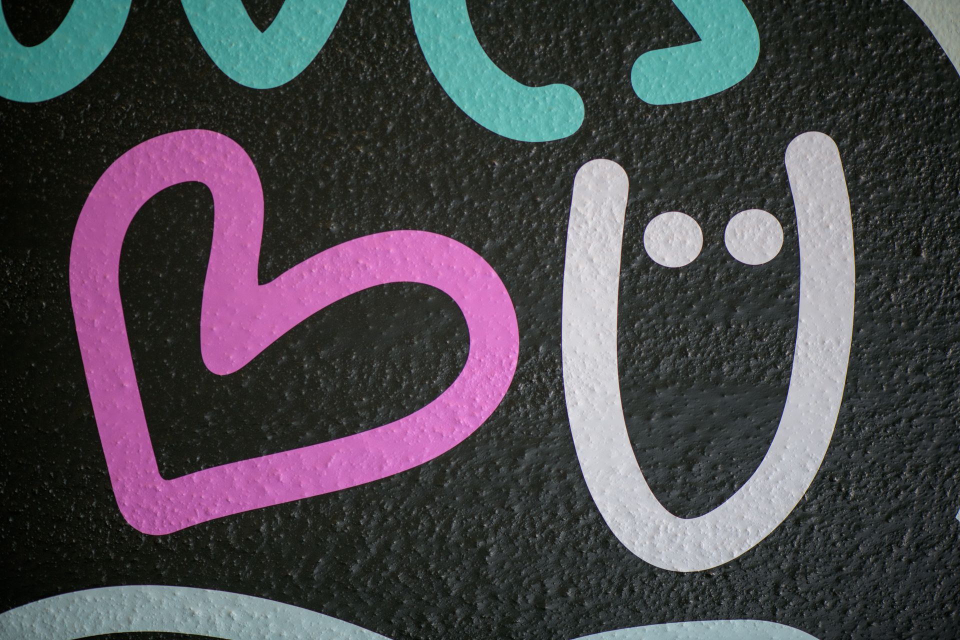 A close-up of a black textured surface featuring a pink heart, a light blue letter, and a white U-shaped figure with dots.