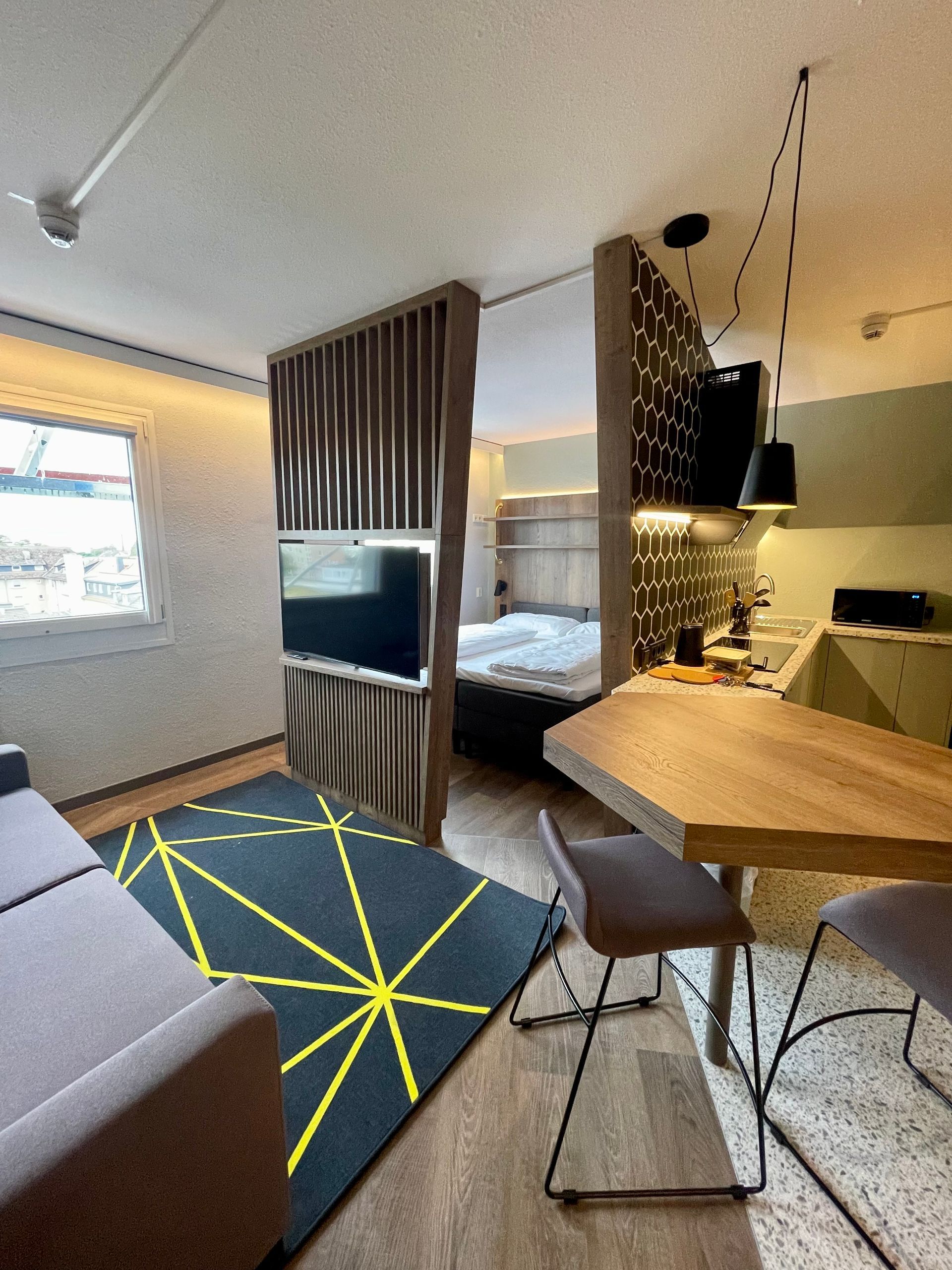 A modern studio apartment interior with a bed, TV partition, rug with geometric yellow lines, and a dining table.