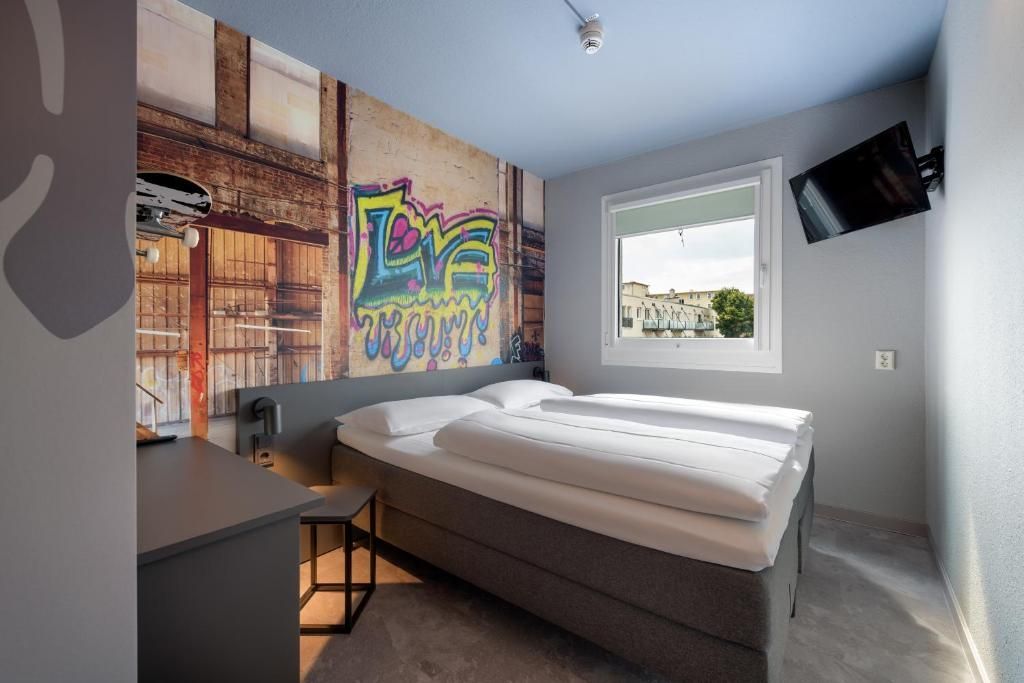 A hotel room with a graffiti-style mural on the headboard wall, a double bed with white bedding, and a wall-mounted TV.