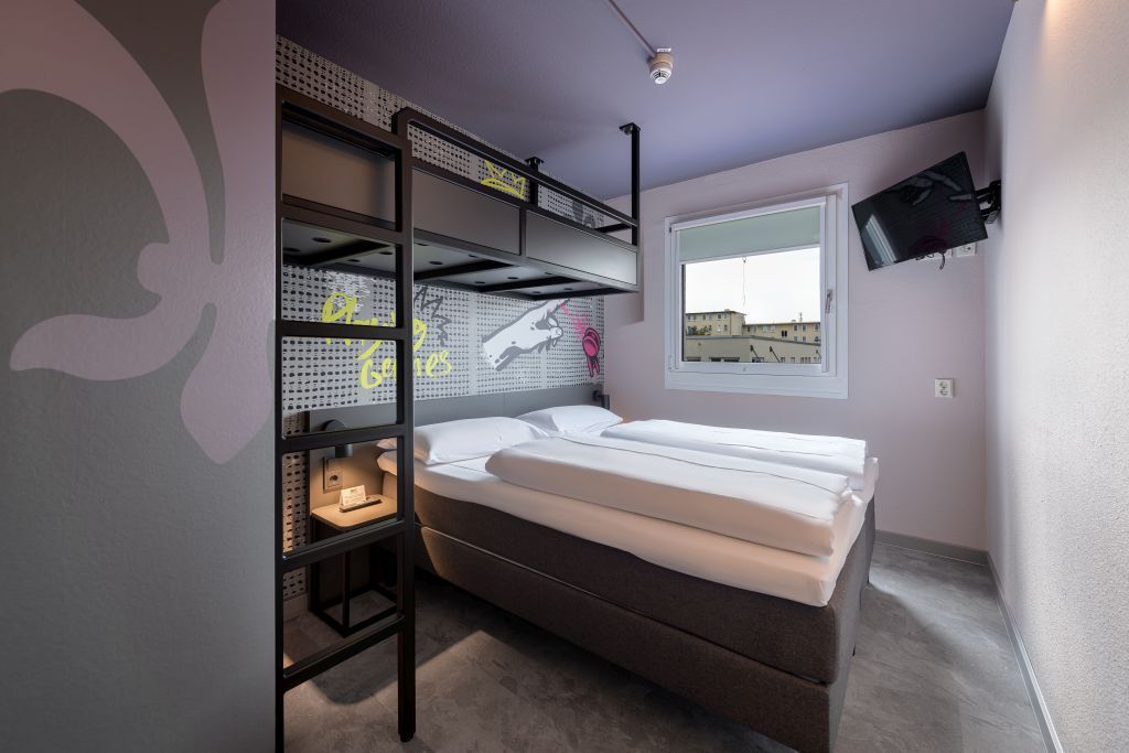 A hotel room with a queen bed, a black metal bunk bed overhead, graffiti wall art, and a wall-mounted TV.