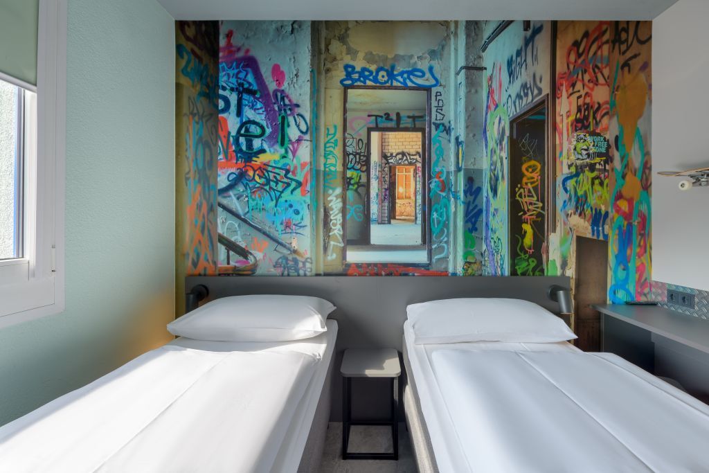 A hotel room featuring two white twin beds separated by a small table against a wall with graffiti-style mural art.