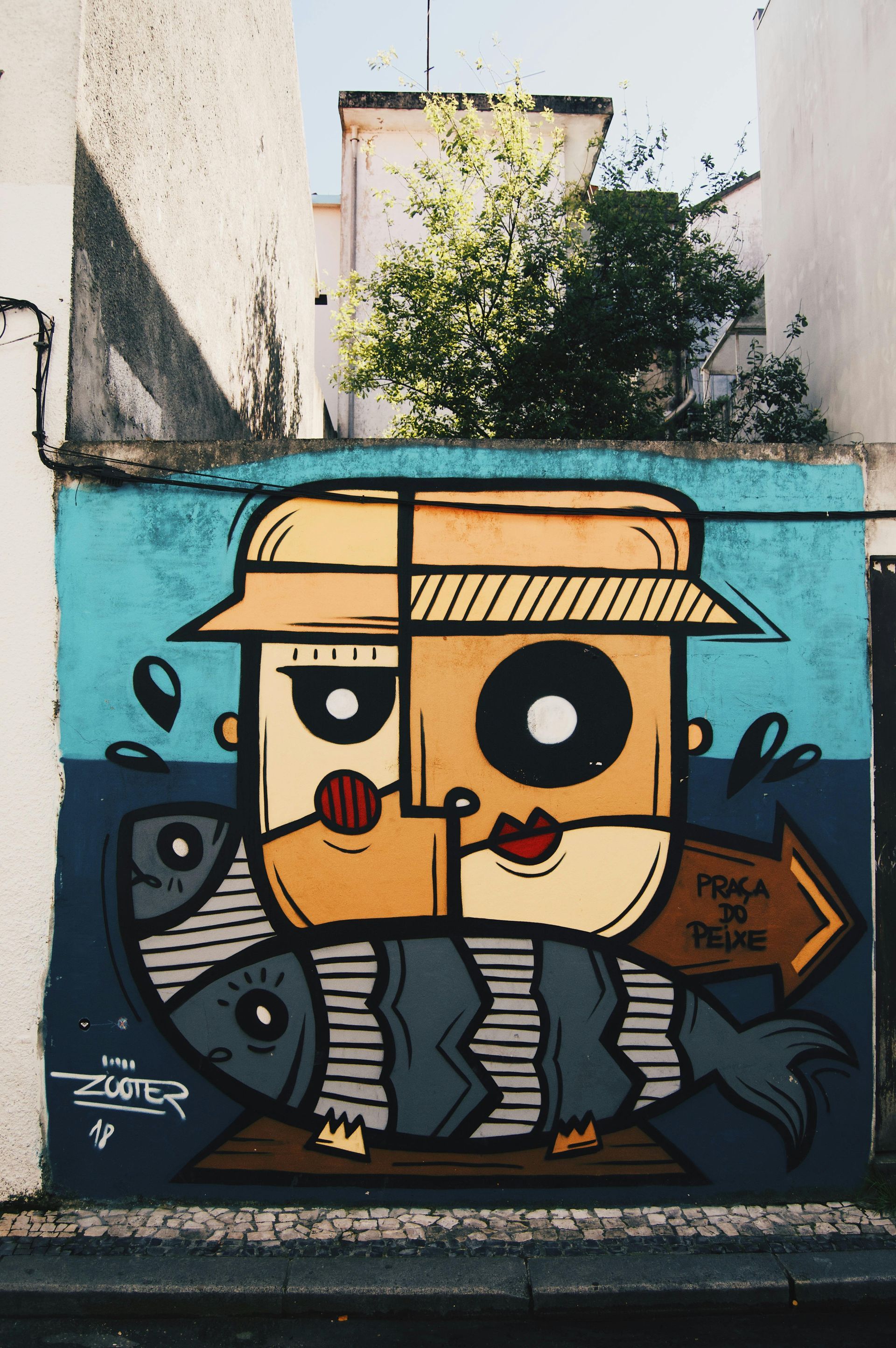 A mural in blue and beige features a stylized face wearing a hat above a striped fish, with a sign pointing to the right.