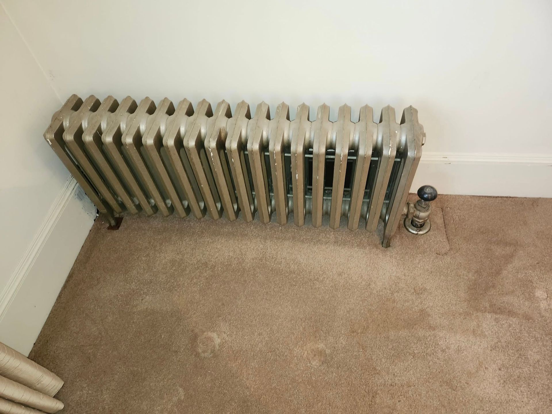 Cast iron radiator in a corner of a room with tan carpet and white walls.