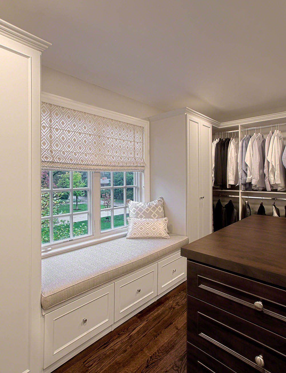 A walk in closet with a window seat and a dresser.