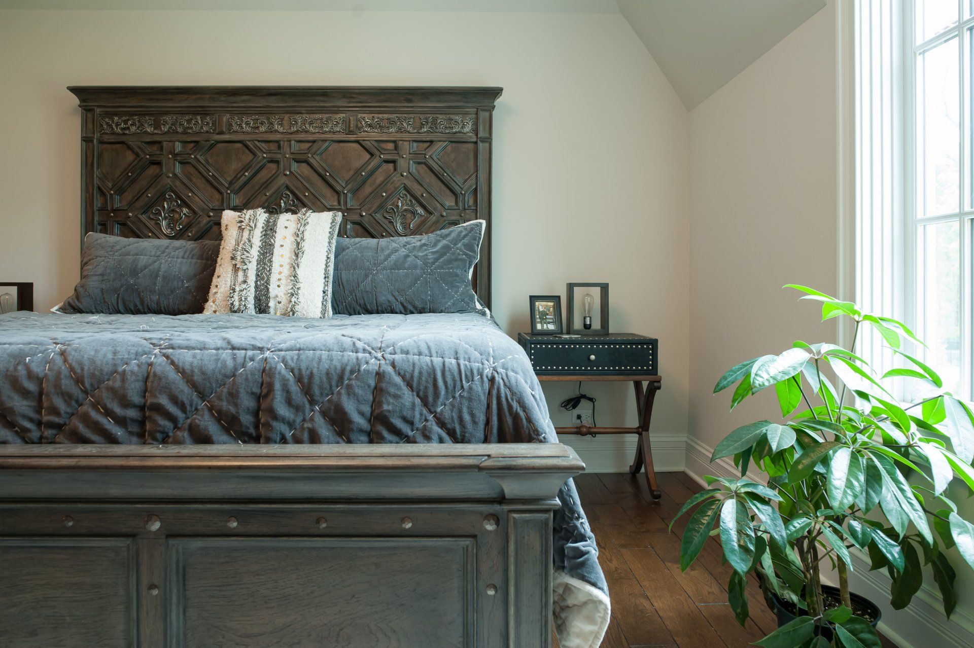 A bedroom with a bed , nightstand , plant and window.