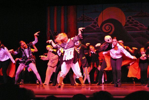 Dancers on stage perform with costumes and a backdrop. Brightly lit with various colors, they gesture and move dynamically.