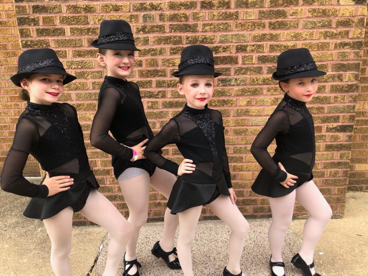 Four young dancers in black costumes and hats pose in front of a brick wall.