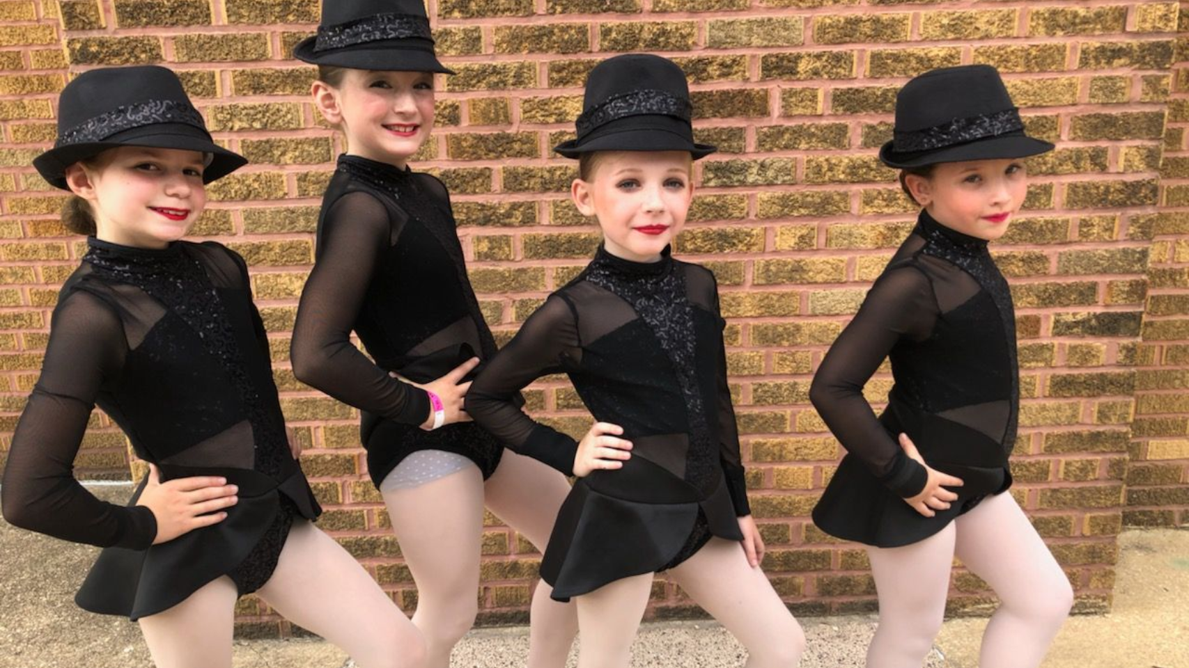 Four young dancers in black costumes and hats pose in front of a brick wall.