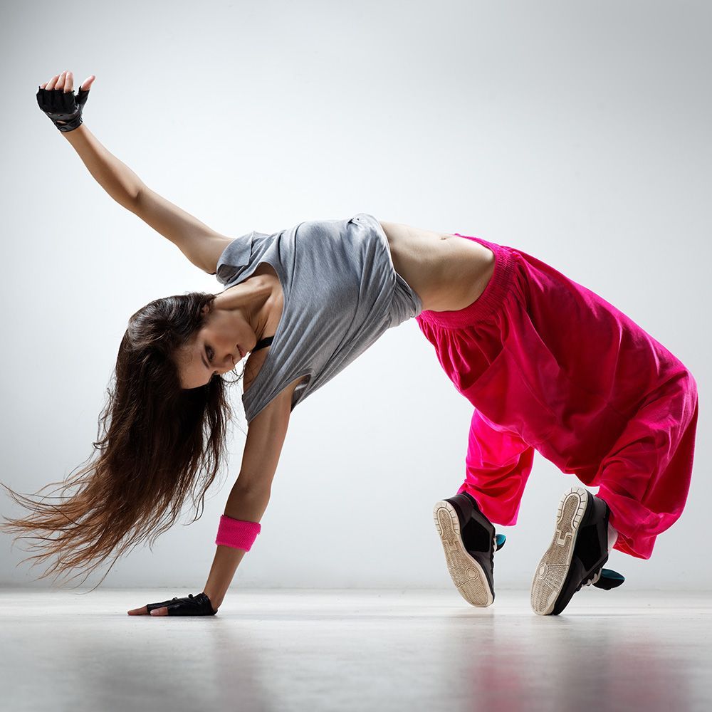 Dancer arched backward, arm raised. Wearing gray top, pink pants, and black gloves. White background.