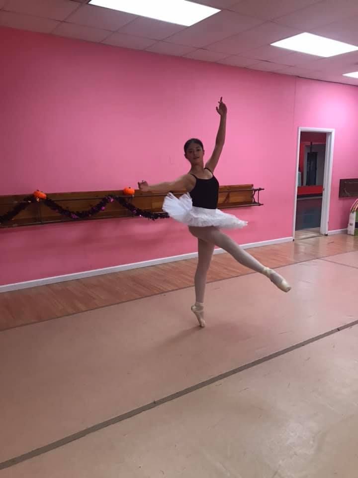 Ballerina in a white tutu, black leotard, and pointe shoes, performing a ballet pose in a pink studio.
