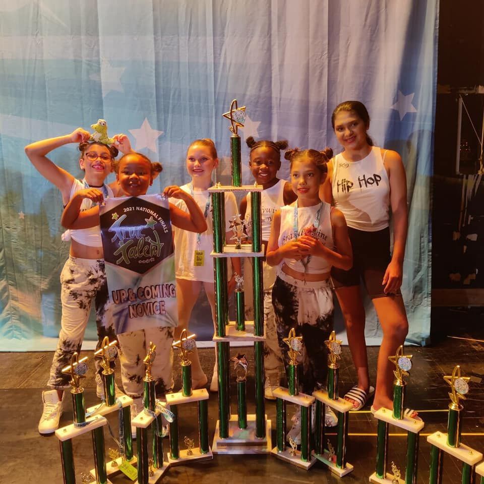 Dancers posing with trophies on a stage with a blue backdrop. They are smiling and holding up awards.