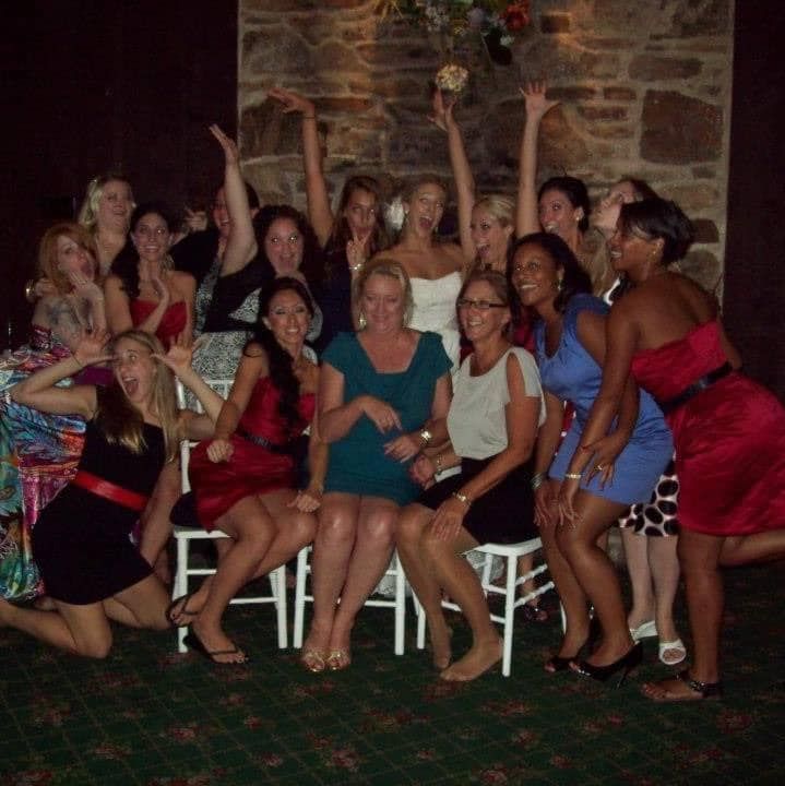 Group of women celebrating. Some seated, others standing, arms raised, smiling, indoors.