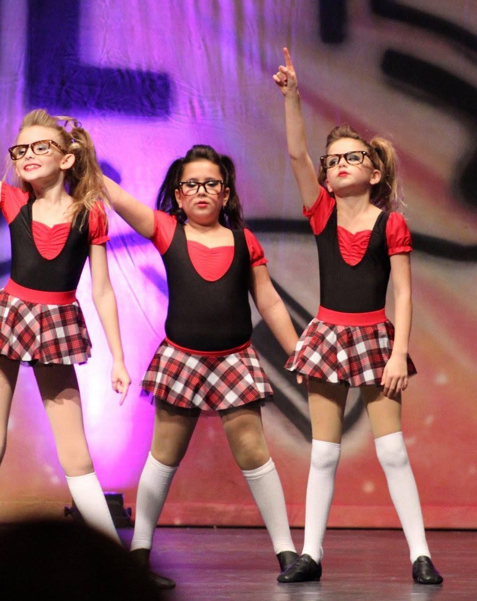 Three young dancers in black tops, plaid skirts, and glasses, performing onstage.