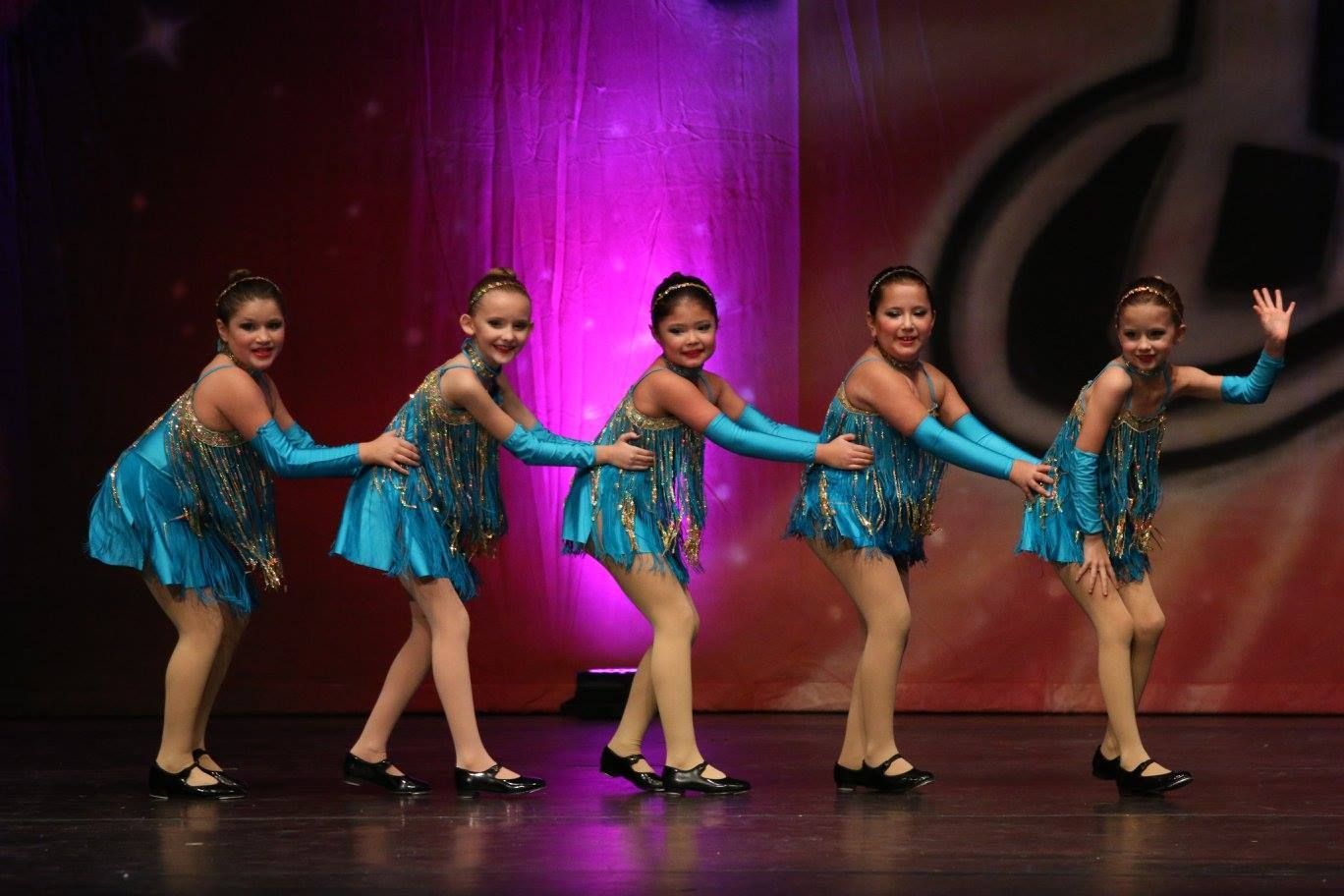 Five dancers in turquoise costumes on a stage, linked together, smiling, with one waving.